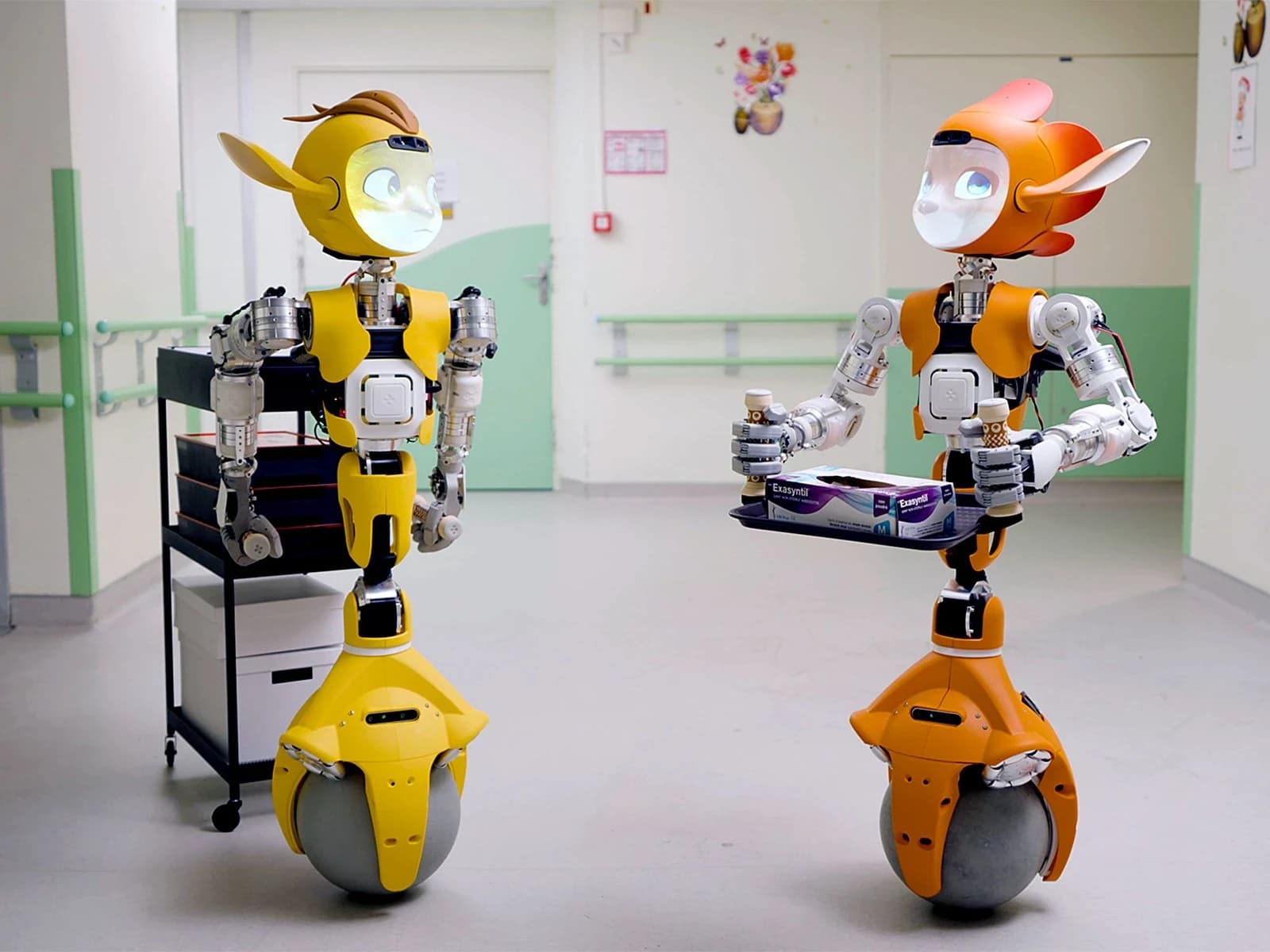 Two Mirokaï robots in a hospital corridor, one carrying a tray of supplies