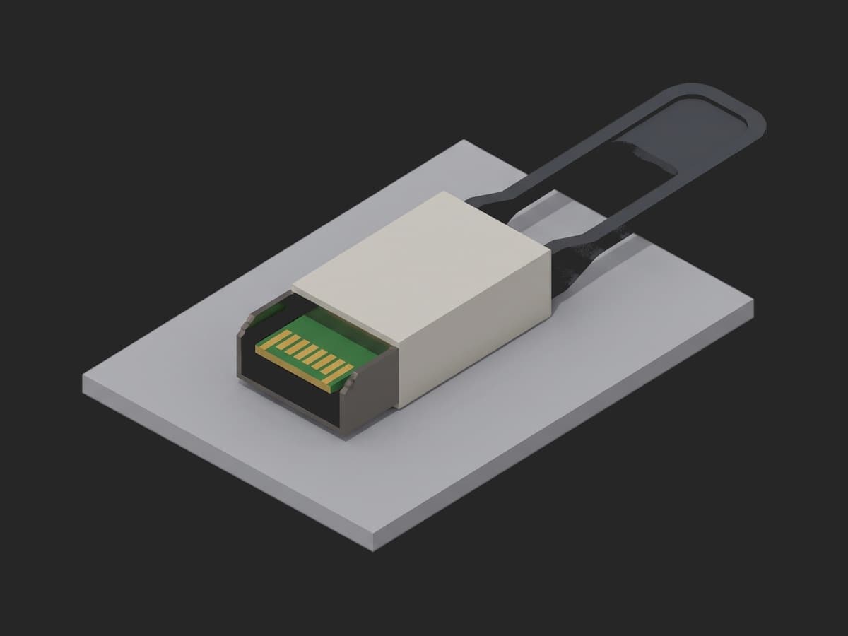 A QSFP-DD loopback plug on a small light-gray plate: an off-white body with a dark-gray pull tab and the gold edge contacts of the green PCB tongue visible at the front.